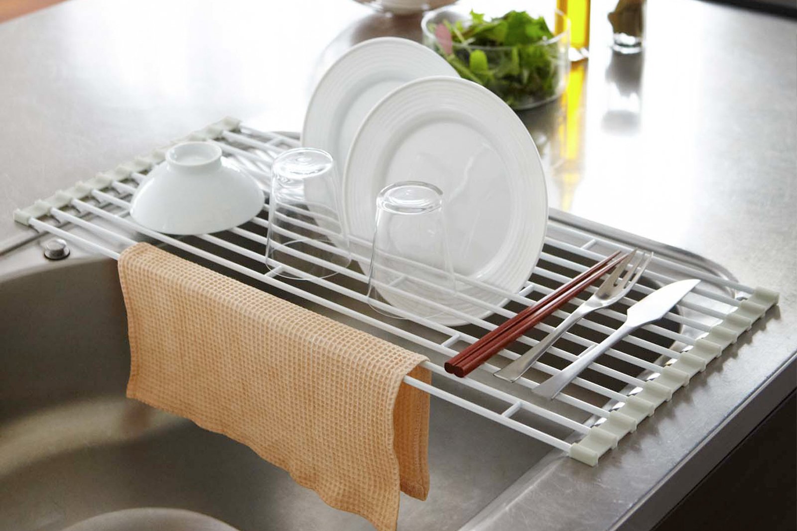 Over-the-Sink Dish Drainer holding plates, cups, bowls, silverware on sink counter by Yamazaki Home.