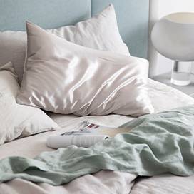 Bed styled with the Smoke Gray Linen Duvet Cover Set, a Smoke Gray Linen Fitted Sheet, Sage Linen Flat Sheet with Border and a Smoke Gray Silk Linen Flip Pillowcase. Also featured is an open magazine positioned atop the sheets.				