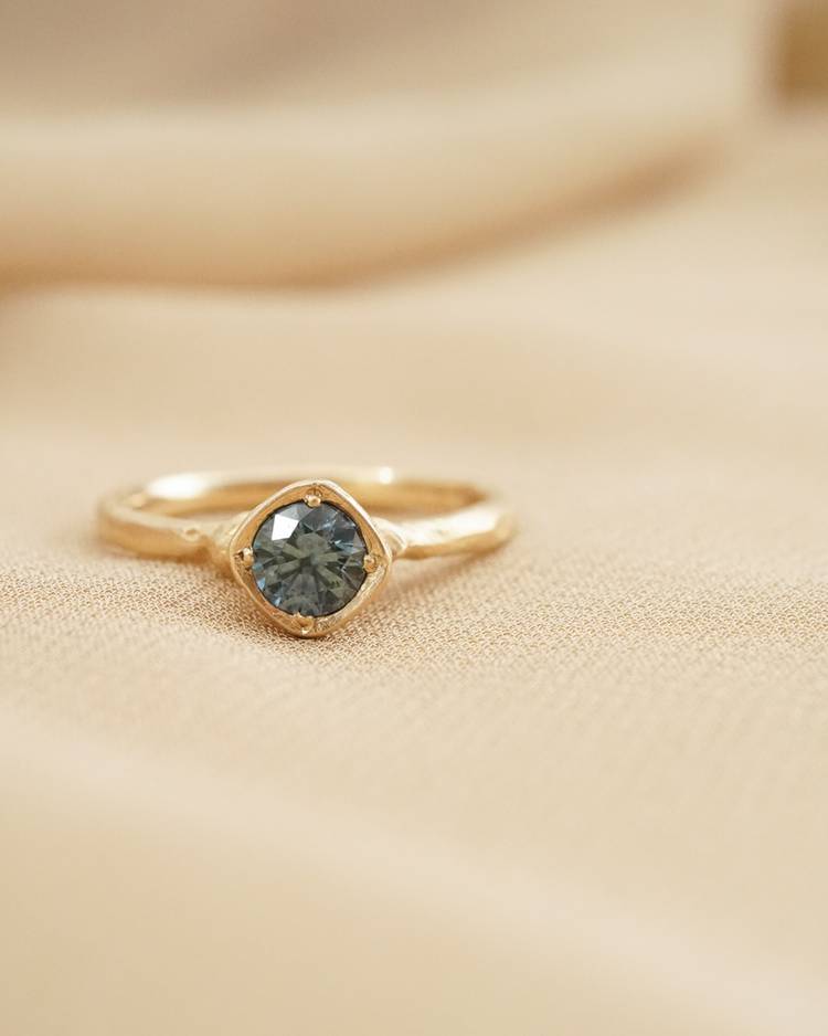 0.5ct Venus Solitaire with Viridian Montana sapphire in 14k recycled gold in a bezel setting with decorative hand carved beads, resting on a tan colored piece of fabric. 