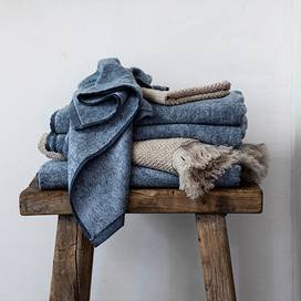 A Denim Bath Towel, Denim Hand Towel, Denim Wash Cloth, Pure Linen Bath Towel and Pure Linen Wash Cloth both in the Natural color sit atop a wooden stool. 
