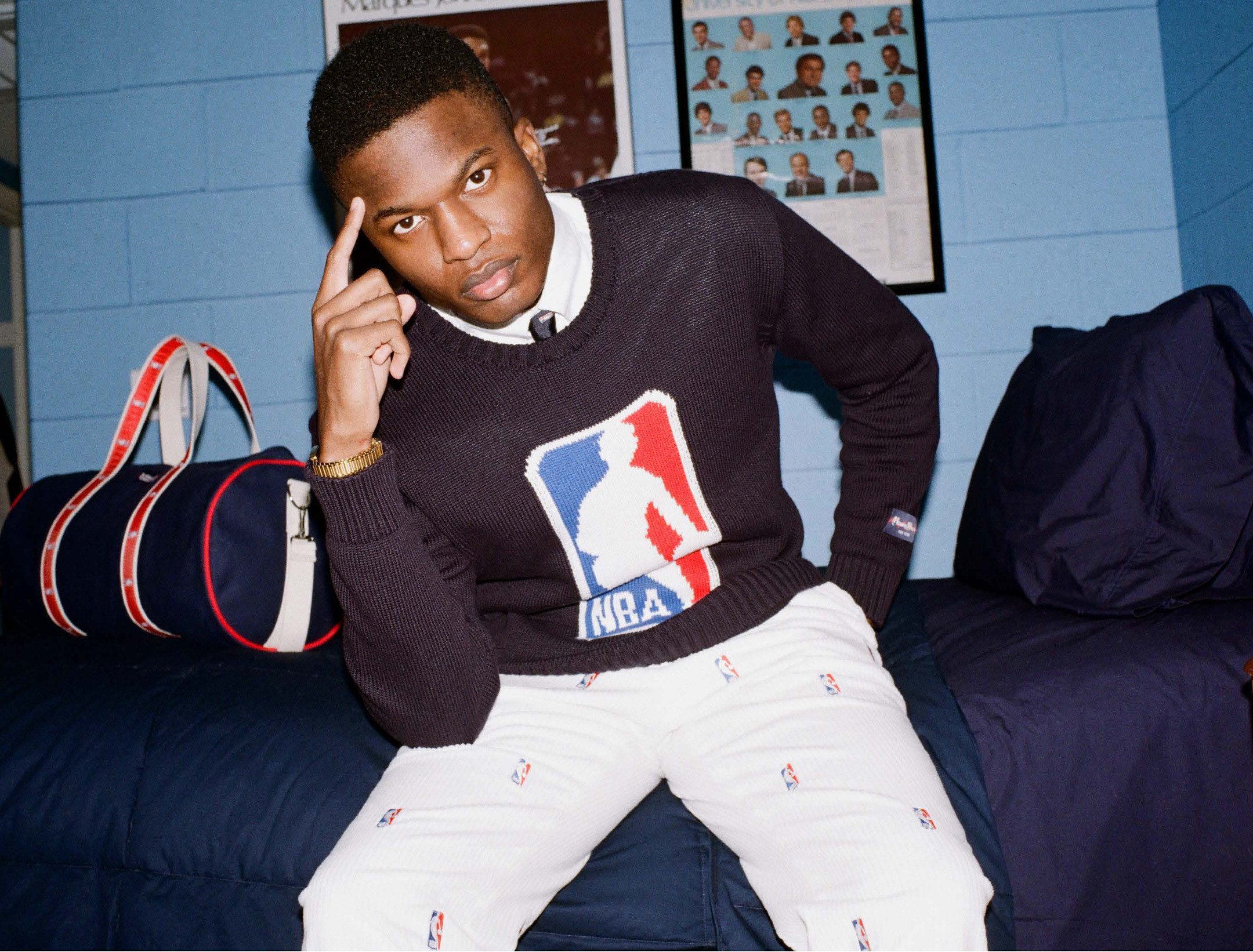 Model wearing the NBA Logo Sweater in Navy on a bed next to the NBA Logo Banker Bag.