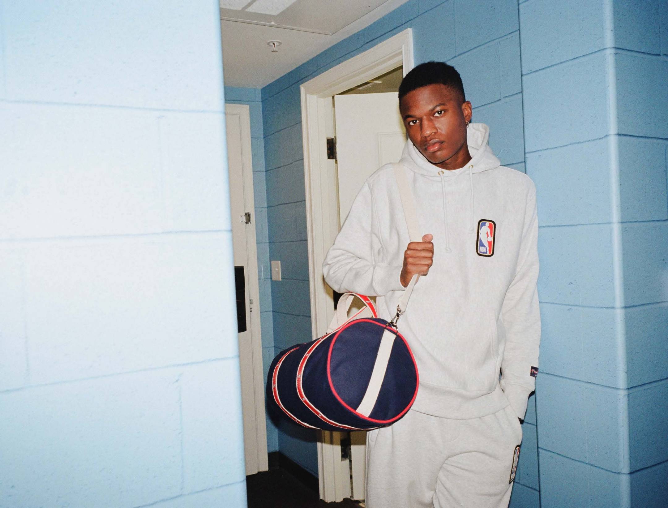 Model in the NBA Logo Hoodie and Joggers holding the NBA Logo Banker Bag in a dorm room.